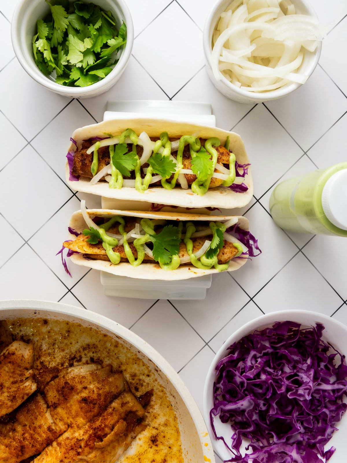 Two fully assembled blackened tilapia tacos with cabbage, fish, onion, cilantro, and avocado sauce, surrounded by ingredients used.