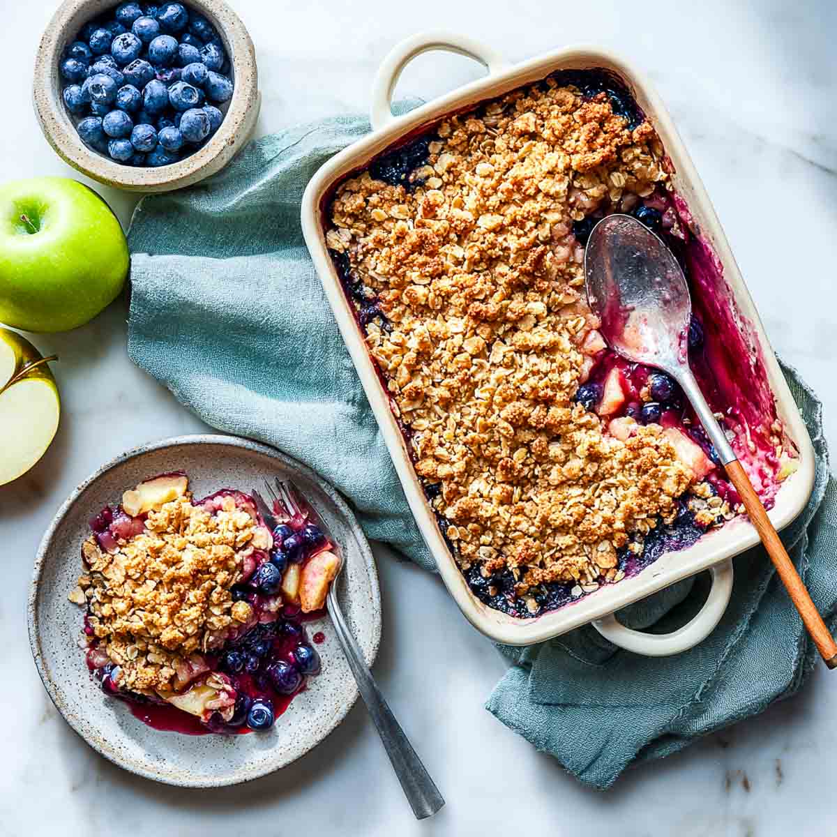 Top-down view of blueberry apple crisp with one portion served on a ceramic plate beside baking dish.