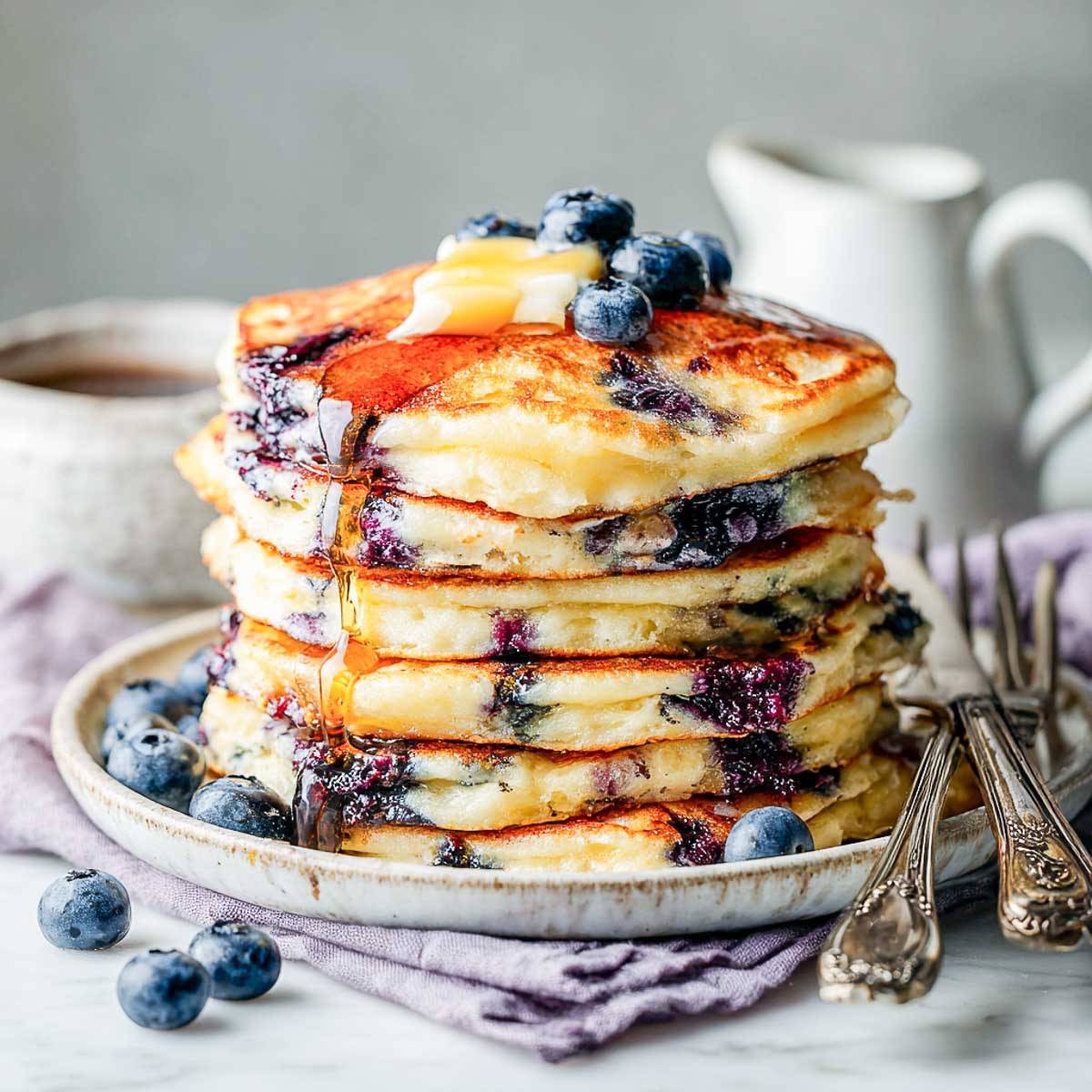 Side view of fluffy blueberry pancakes stacked with syrup, butter, and fresh berries.