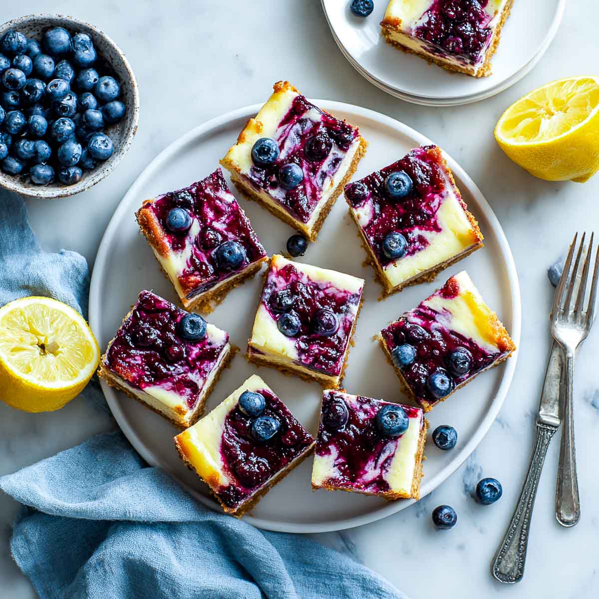 Top-down view of blueberry lemon cheesecake bars cut into squares with fresh blueberries and lemon halves.
