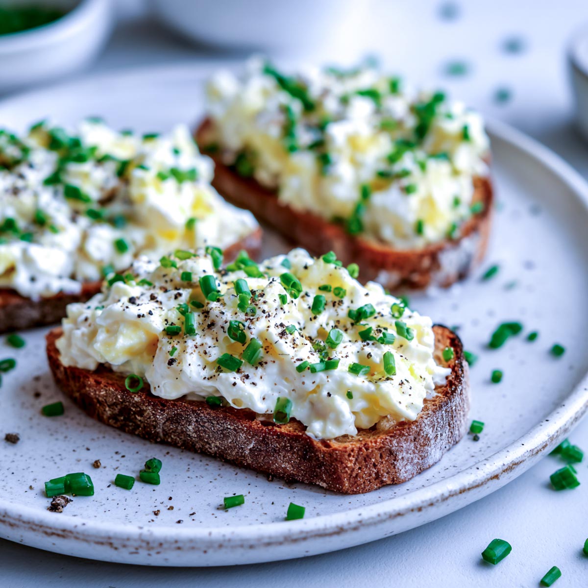 Close-up of cottage cheese egg salad spread on slices of whole-grain toast, topped with fresh chives and cracked black pepper, arranged on a white ceramic plate for an inviting serving suggestion.