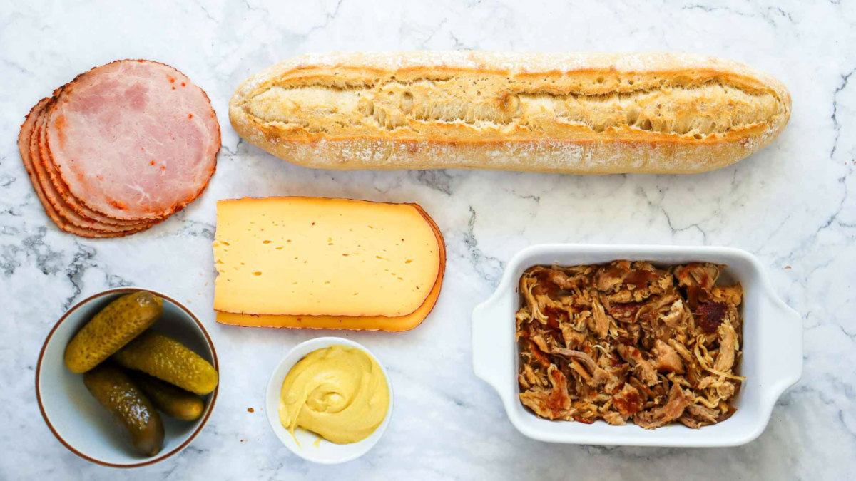 Ingredients for a Cuban sandwich including ham, Swiss cheese, mustard, pickles, shredded pork, and a loaf of bread on a marble surface.