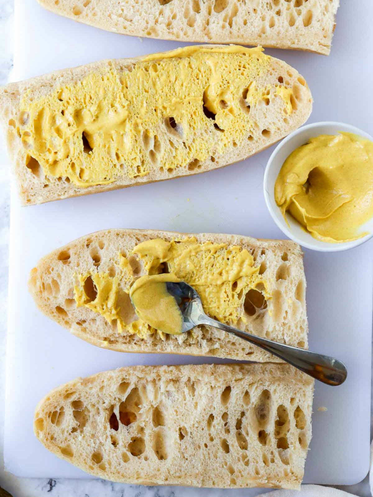 Bread slices spread with yellow mustard on a cutting board with a spoon and bowl.