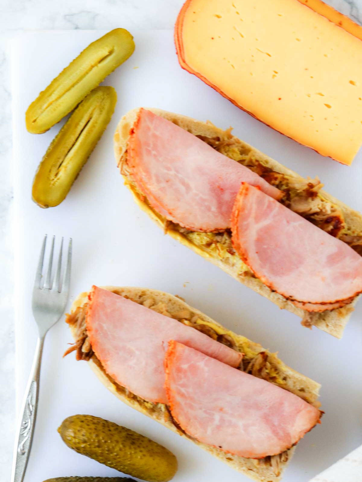 Layered ham and pulled pork on sandwich bread with cheese and pickles in the background.