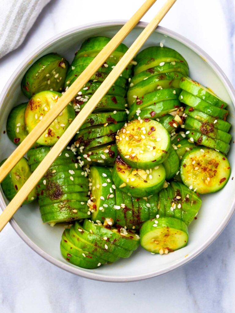 Top-down view of Korean cucumber salad served in a white bowl, drizzled with spicy chili oil dressing and sprinkled with sesame seeds, with chopsticks resting on top.