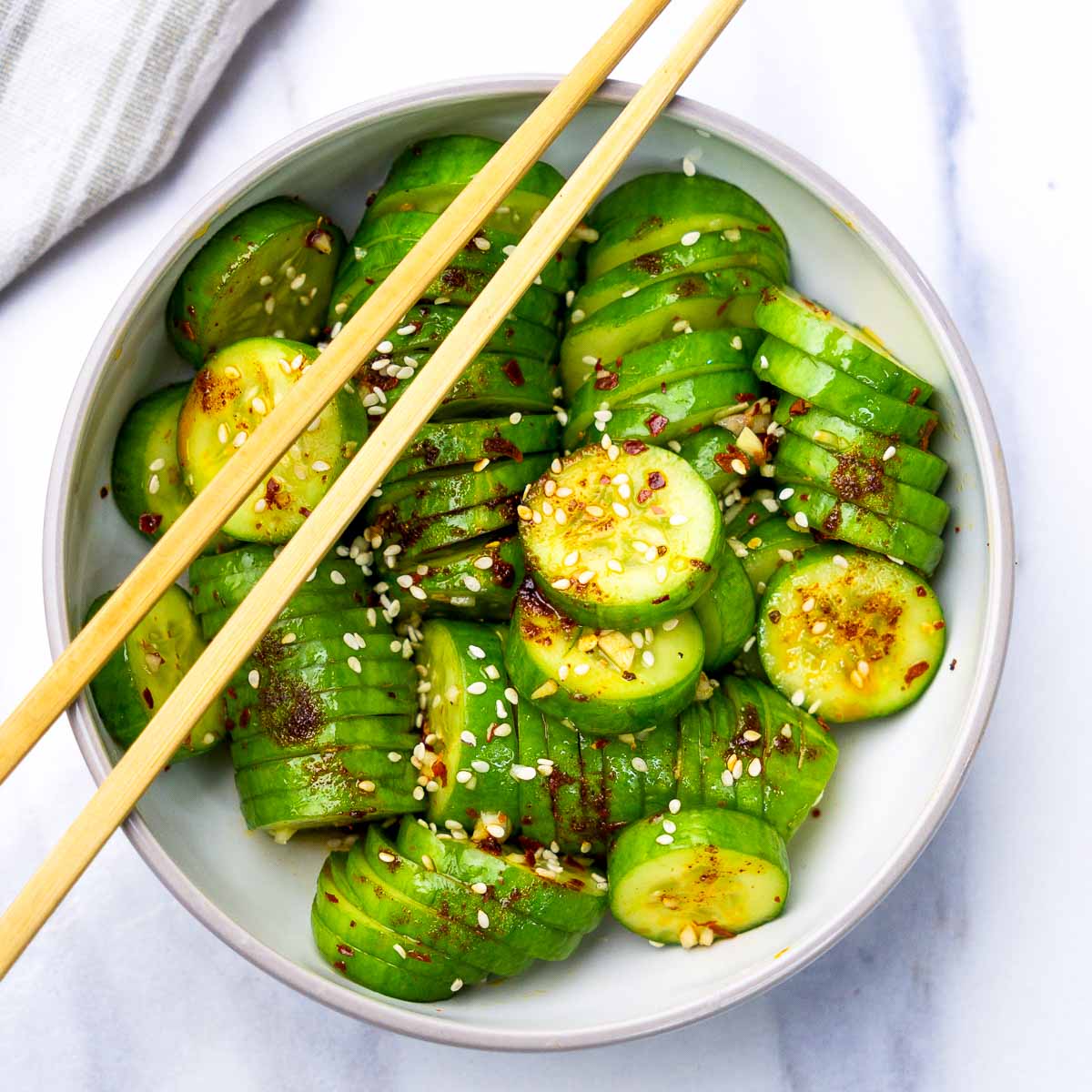 Flat lay of accordion-sliced cucumber salad with garlic chili oil, sesame seeds, and soy-based dressing, served in a bowl with chopsticks.