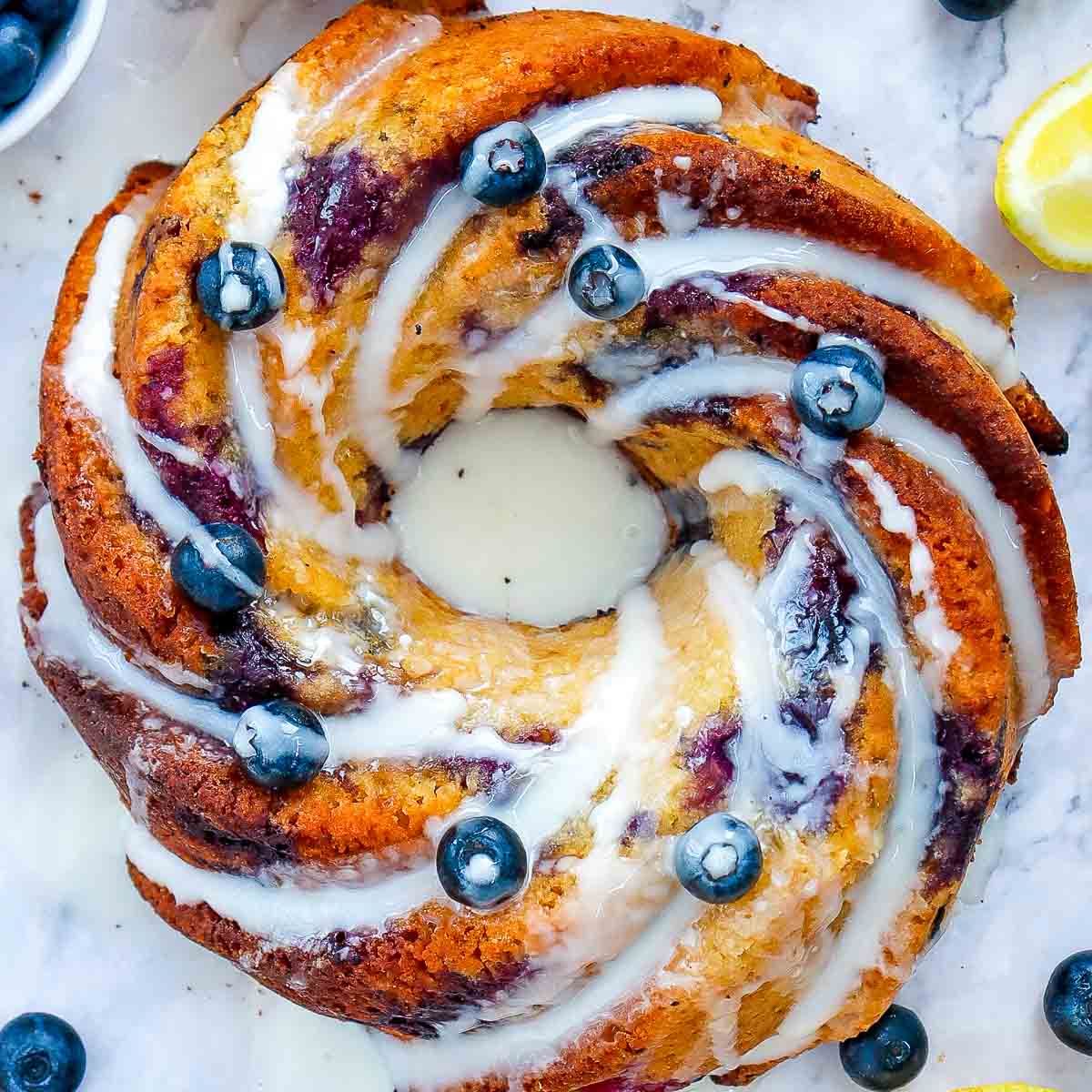 Top-down view of a lemon blueberry bundt cake with a golden crust, drizzled with lemon glaze and decorated with fresh blueberries on a marble background.