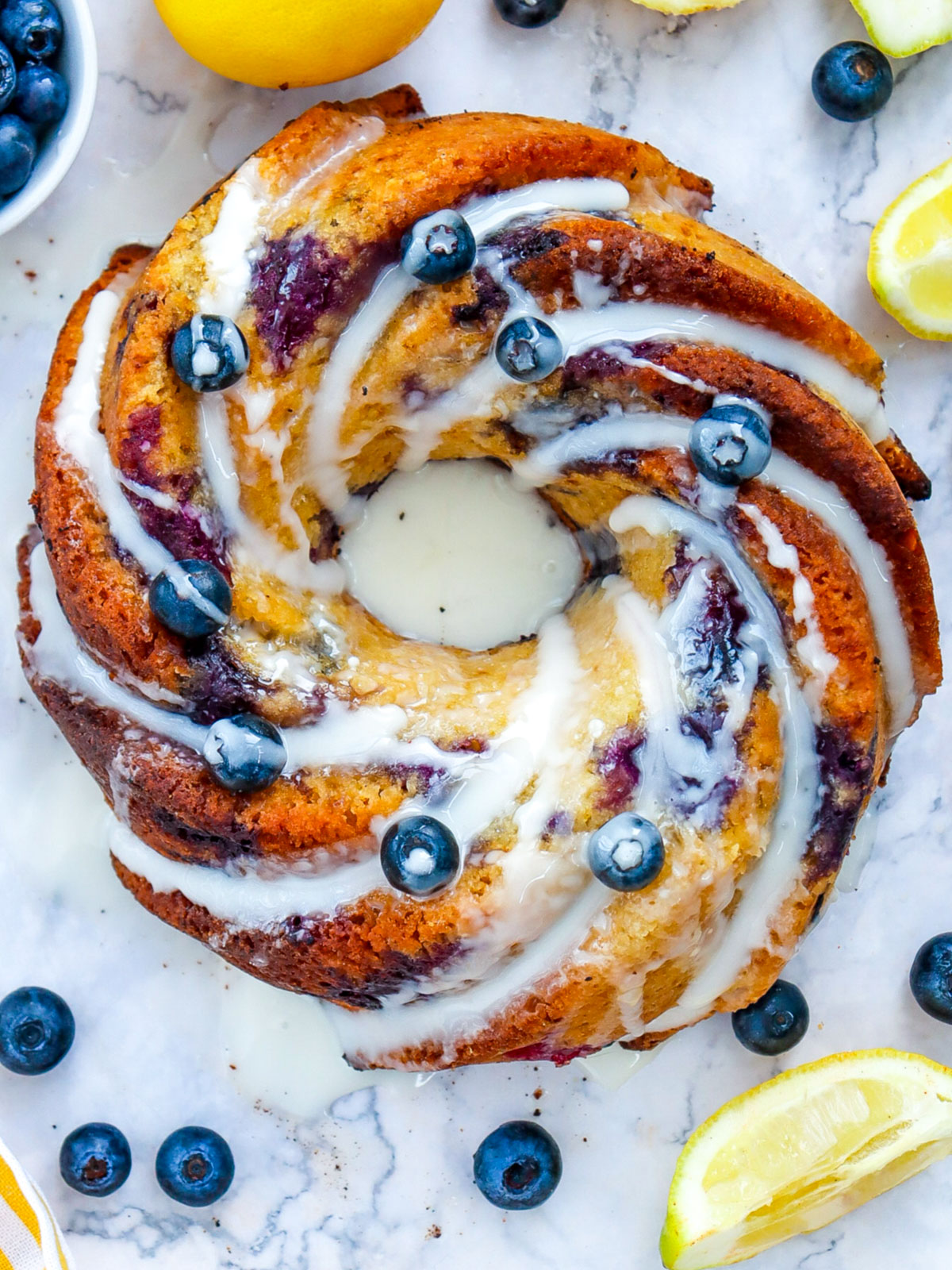 Overhead shot of a glazed lemon blueberry bundt cake surrounded by lemons and blueberries, showcasing the cake’s spiral design and juicy berry swirls.