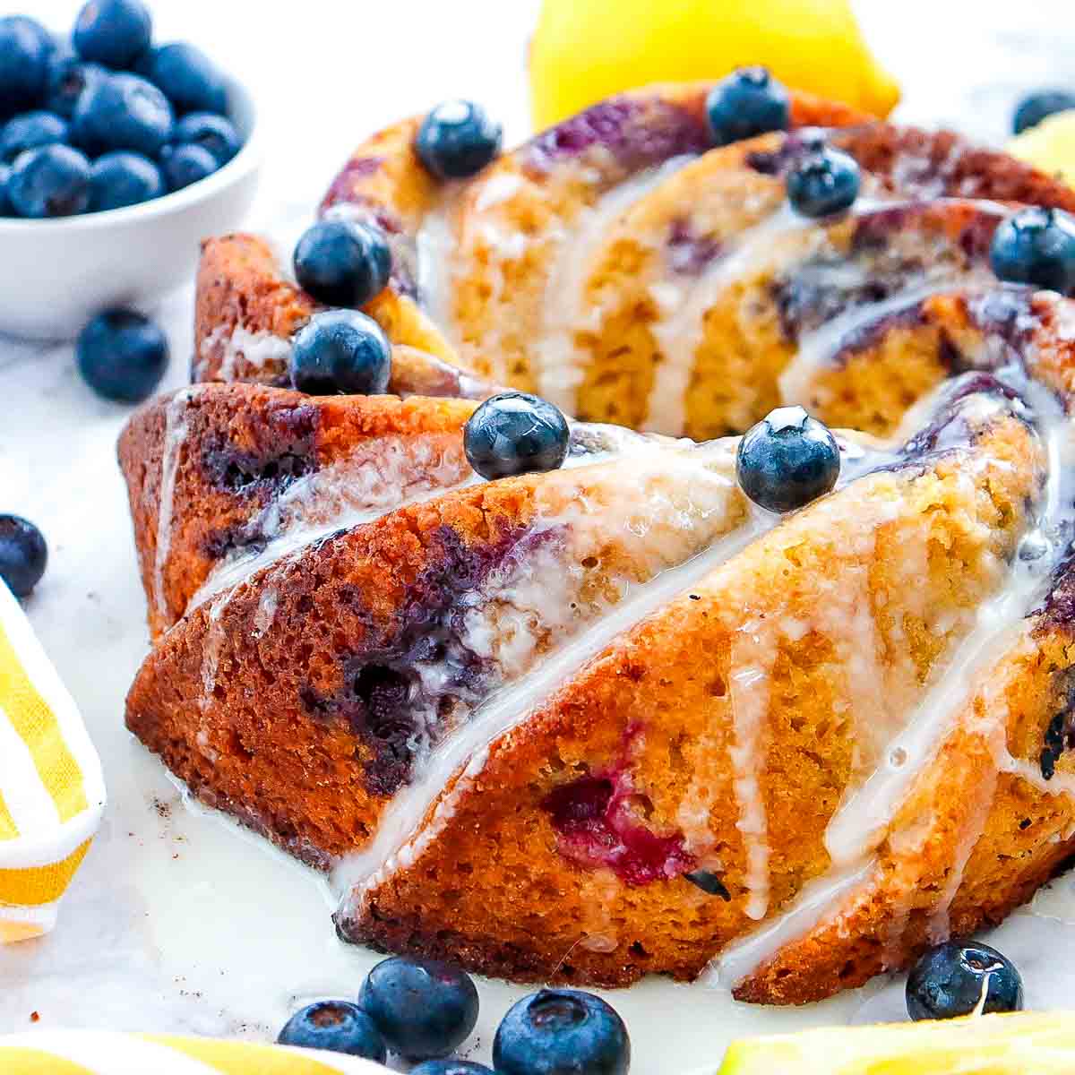Angled side view of a golden lemon blueberry bundt cake with a thick glaze and whole blueberries on top, served on a white marble surface.