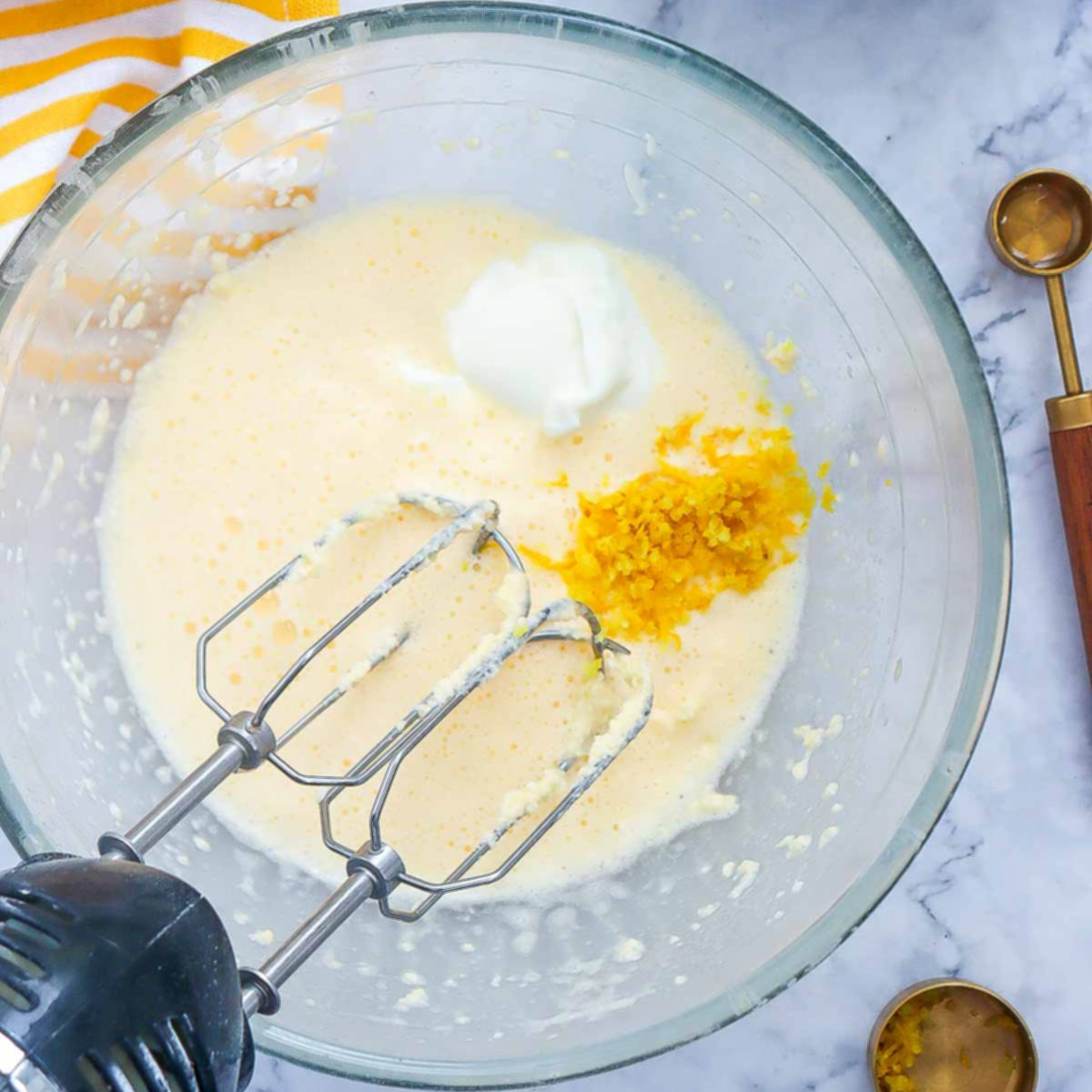 Whipped eggs and butter mixture in a glass bowl with lemon zest and Greek yogurt added, ready to be blended.