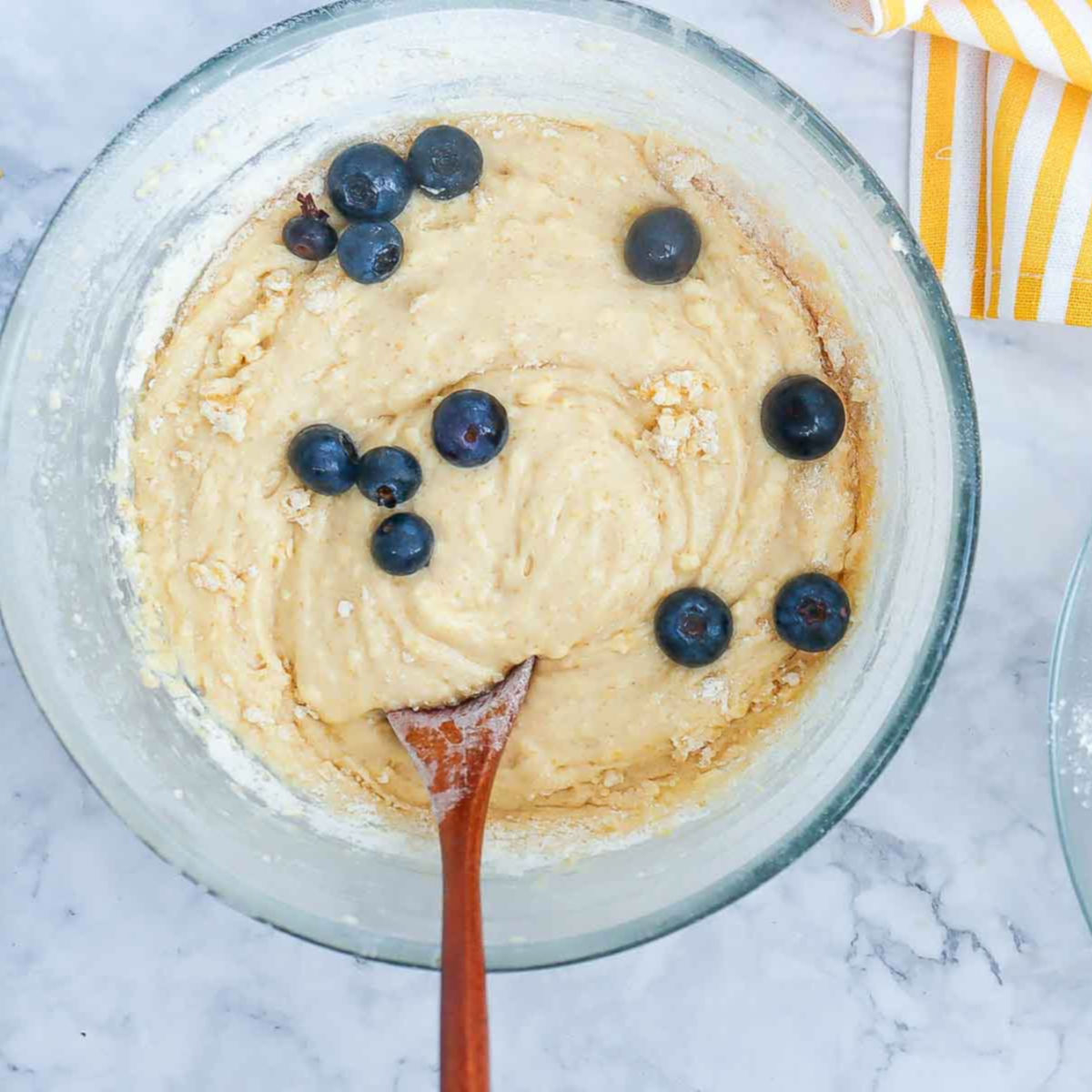 Blueberries being folded into the lemon cake batter in a large mixing bowl using a wooden spoon.