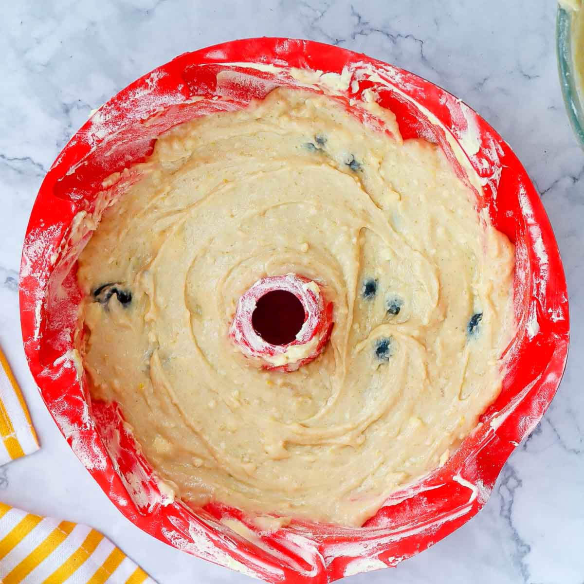 Lemon blueberry cake batter spread evenly in a red bundt pan, ready to go into the oven.