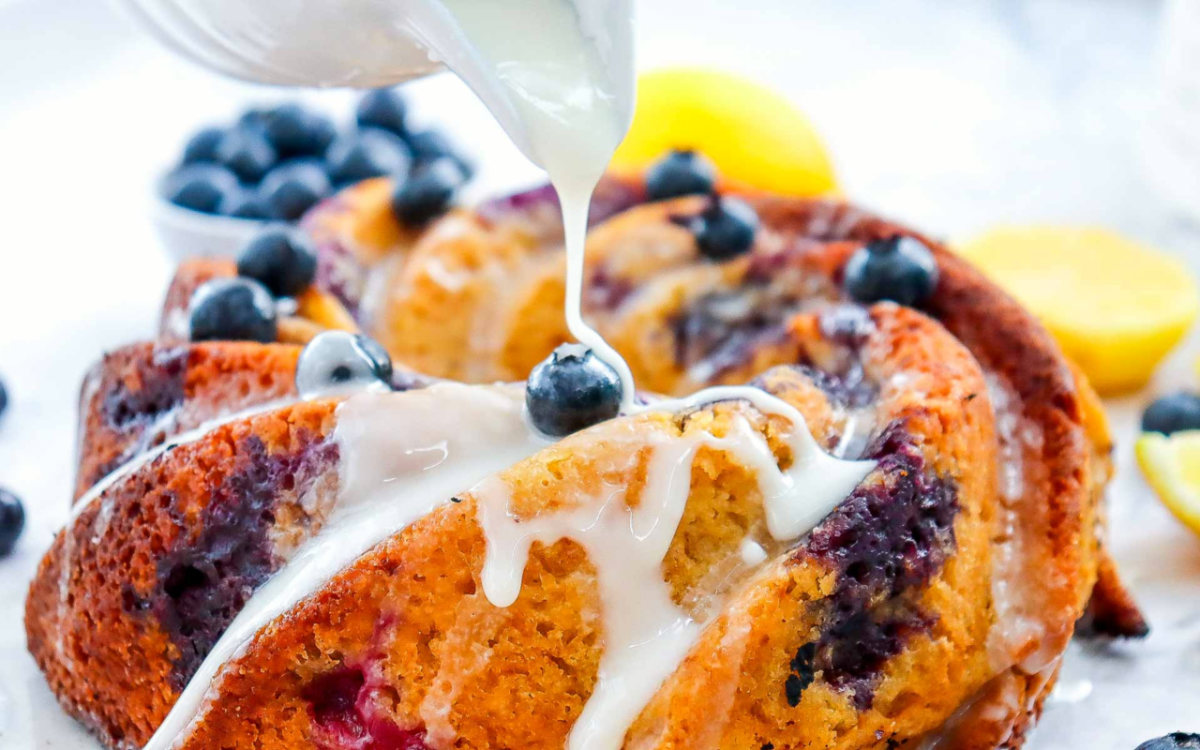 Close-up of lemon glaze being poured over the cooled bundt cake, with blueberries and lemons in the background.