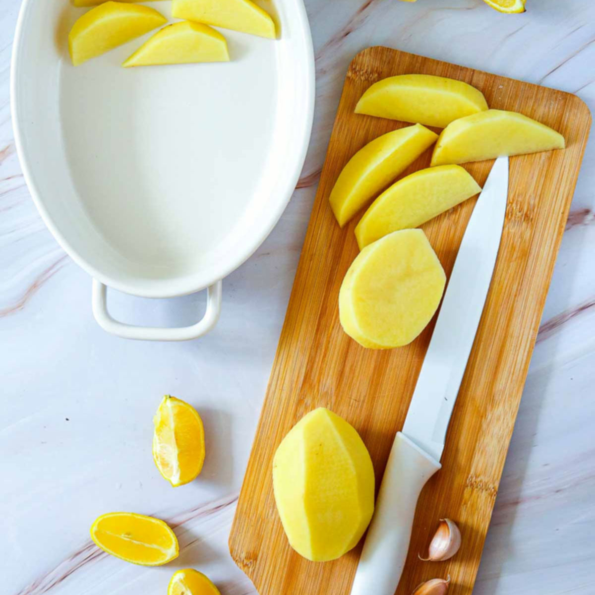 Peeled potatoes being sliced into wedges on a wooden cutting board next to a white baking dish and lemon wedges.
