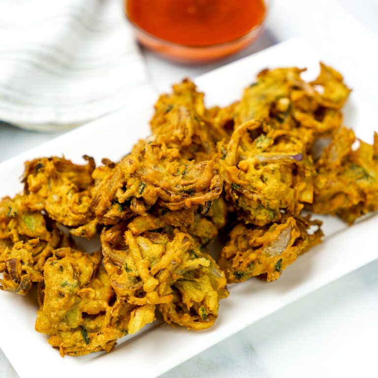 Crispy onion pakoras served on a white rectangular plate with a bowl of red chutney in the background.