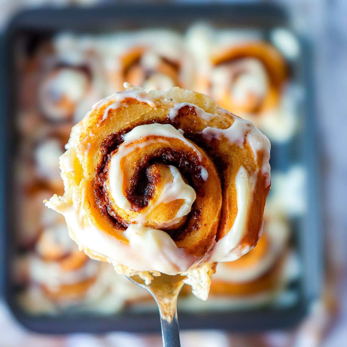Close-up of a single pumpkin cinnamon roll on a fork, topped with maple cream cheese glaze with pan of rolls blurred in the background.