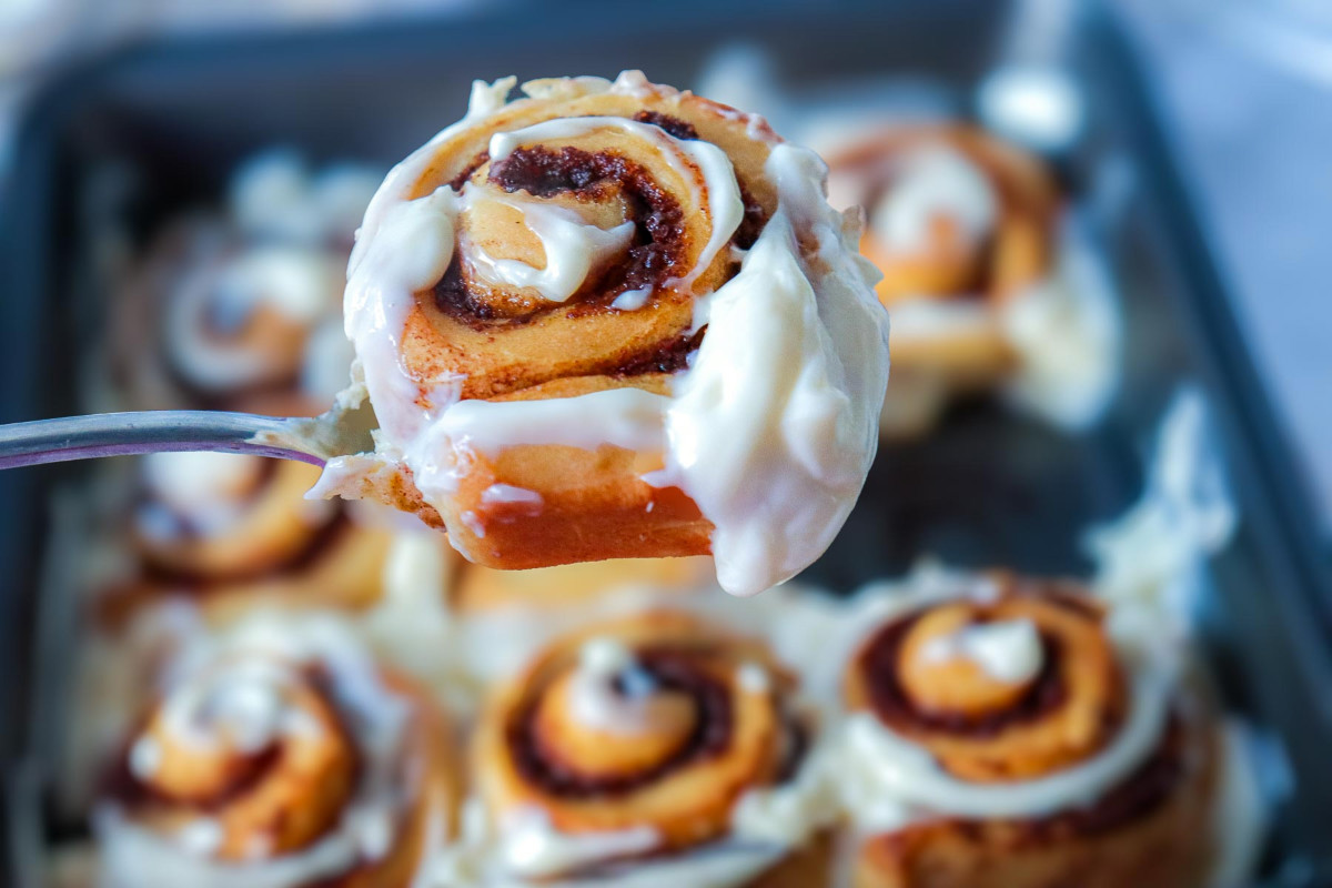 Close-up of a single glazed pumpkin cinnamon roll on a spoon lifted from the pan, showing creamy maple frosting detail.