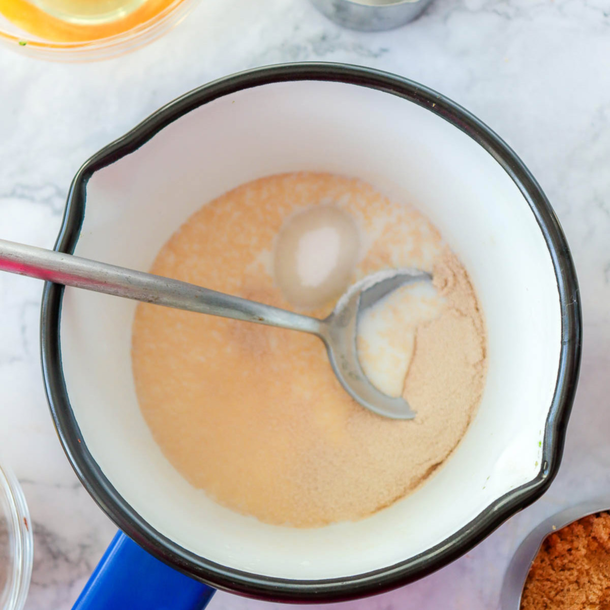 Close-up of warm milk and yeast mixture in a small saucepan with a spoon, showing foamy texture after blooming.