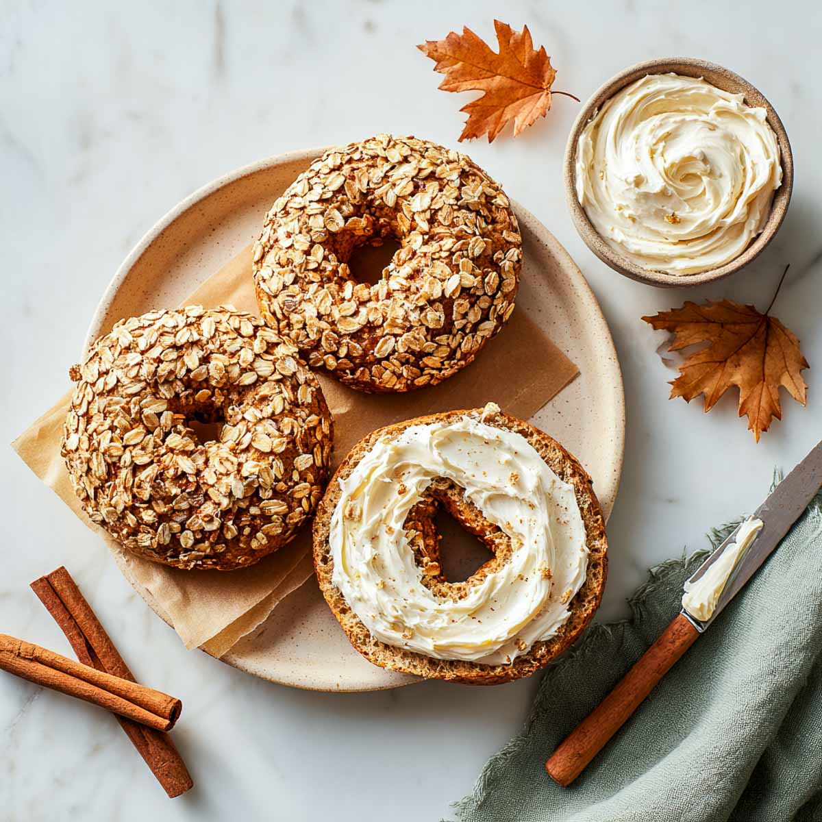 Top-down shot of pumpkin spice bagels with oat topping, one halved and generously spread with maple cream cheese, next to a bowl of whipped spread.