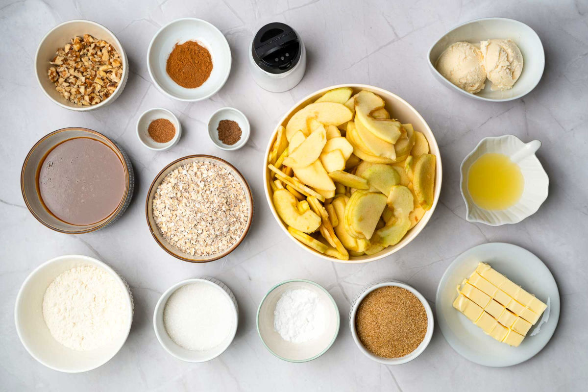 Overhead view of apple crisp ingredients including sliced apples, oats, flour, sugar, butter, spices, nuts, and caramel sauce.