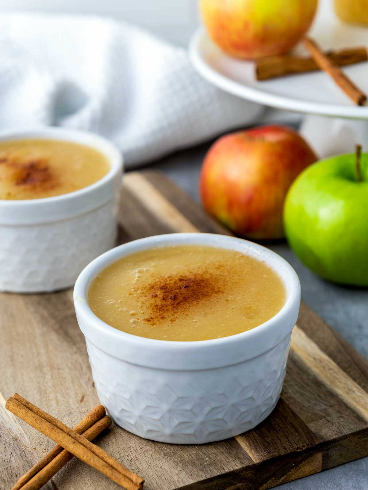 Two small bowls of Instant Pot applesauce with cinnamon, fresh apples, and cinnamon sticks.