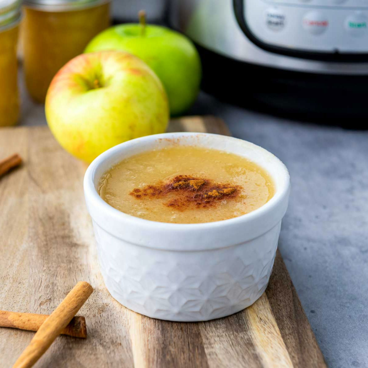 White ramekin of Instant Pot applesauce with apples and jars of homemade applesauce in the background.