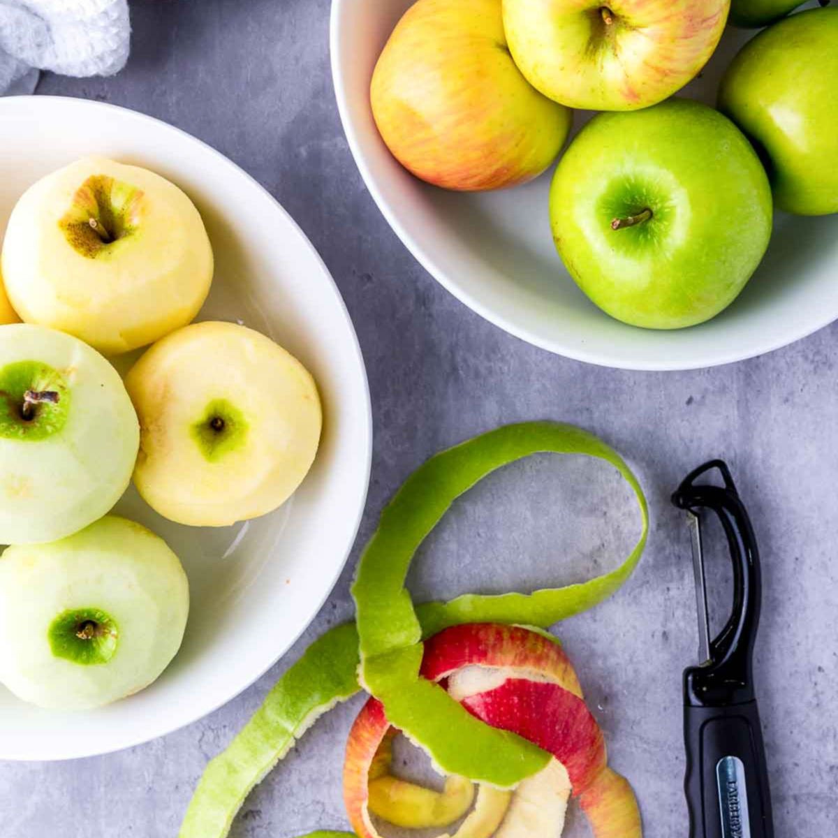 Peeled apples in a bowl with apple peels and a peeler on the side for making applesauce.