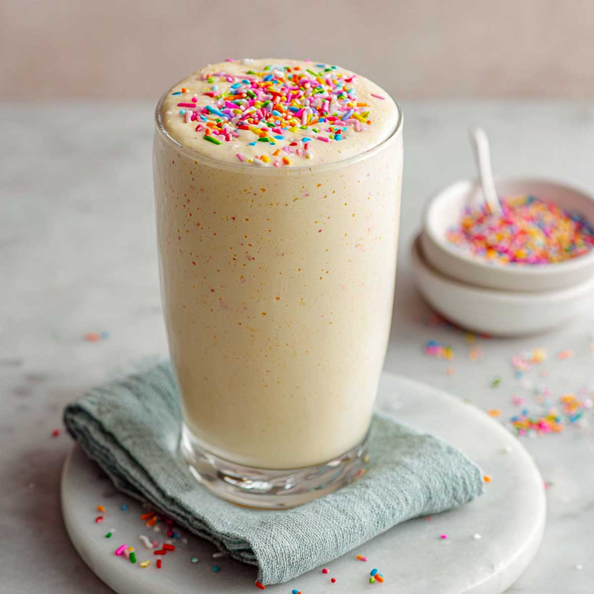 Creamy cake batter protein shake topped with rainbow sprinkles on marble surface.