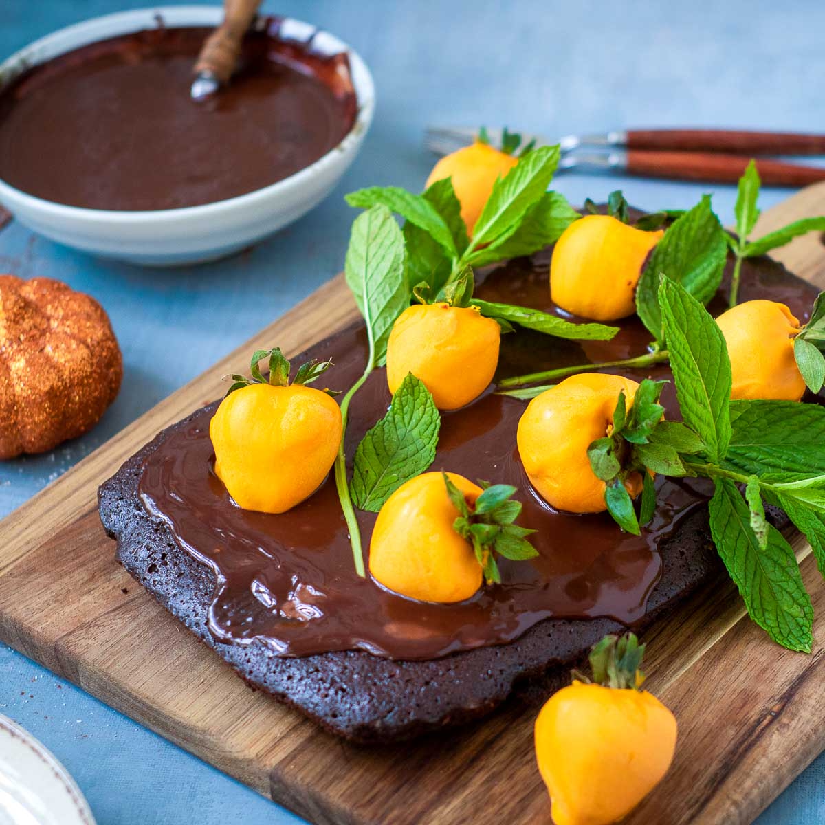 Side view of chocolate brownie cake with glossy ganache, orange-dipped strawberries, and mint leaves on a wooden board.
