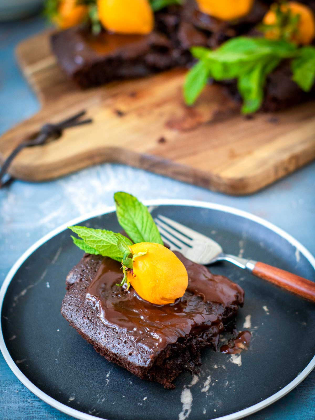 Single square of chocolate brownie cake with ganache, an orange-dipped strawberry pumpkin, and a mint leaf on a black plate.