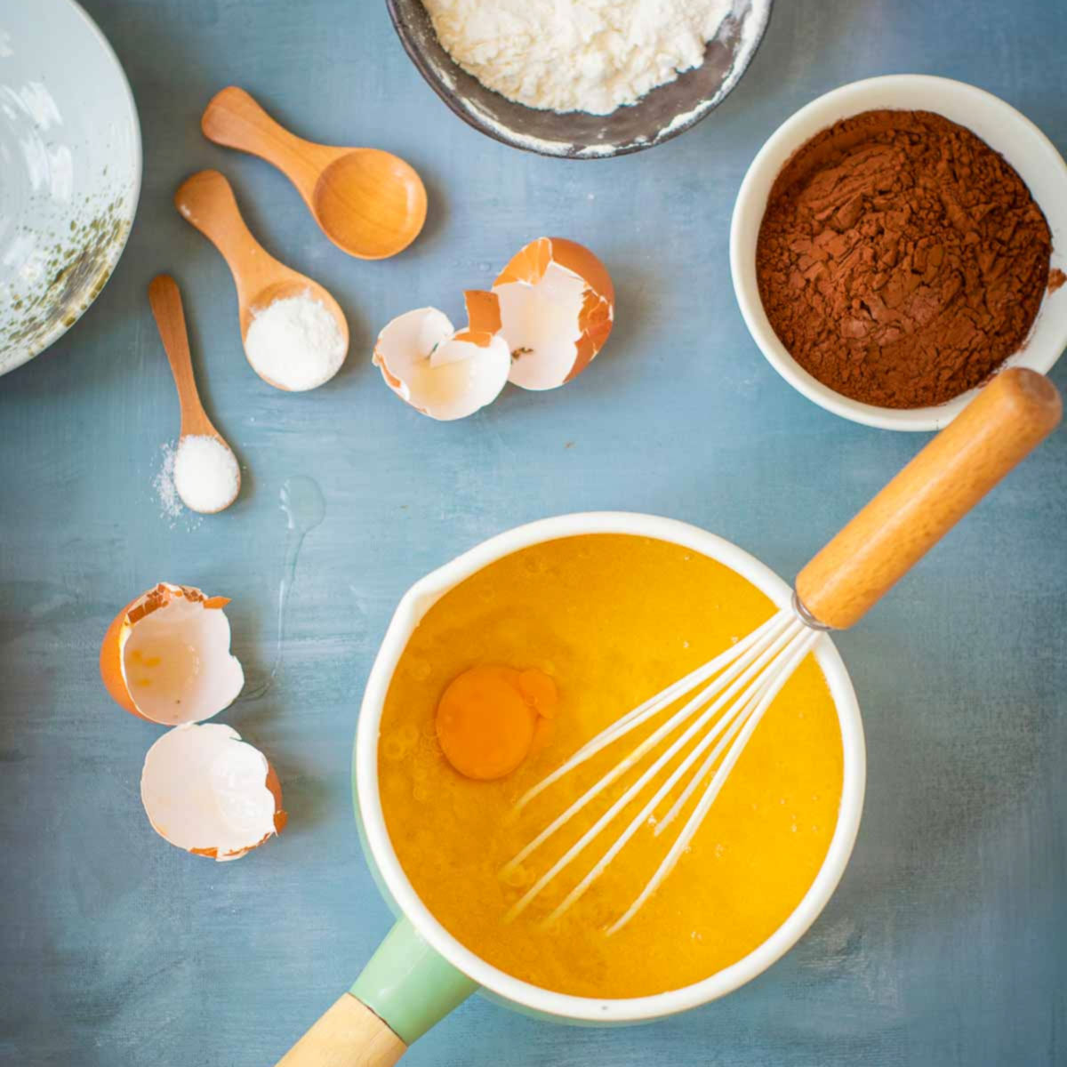 Whisking eggs into melted butter and sugar in a saucepan with baking ingredients arranged on a blue surface.