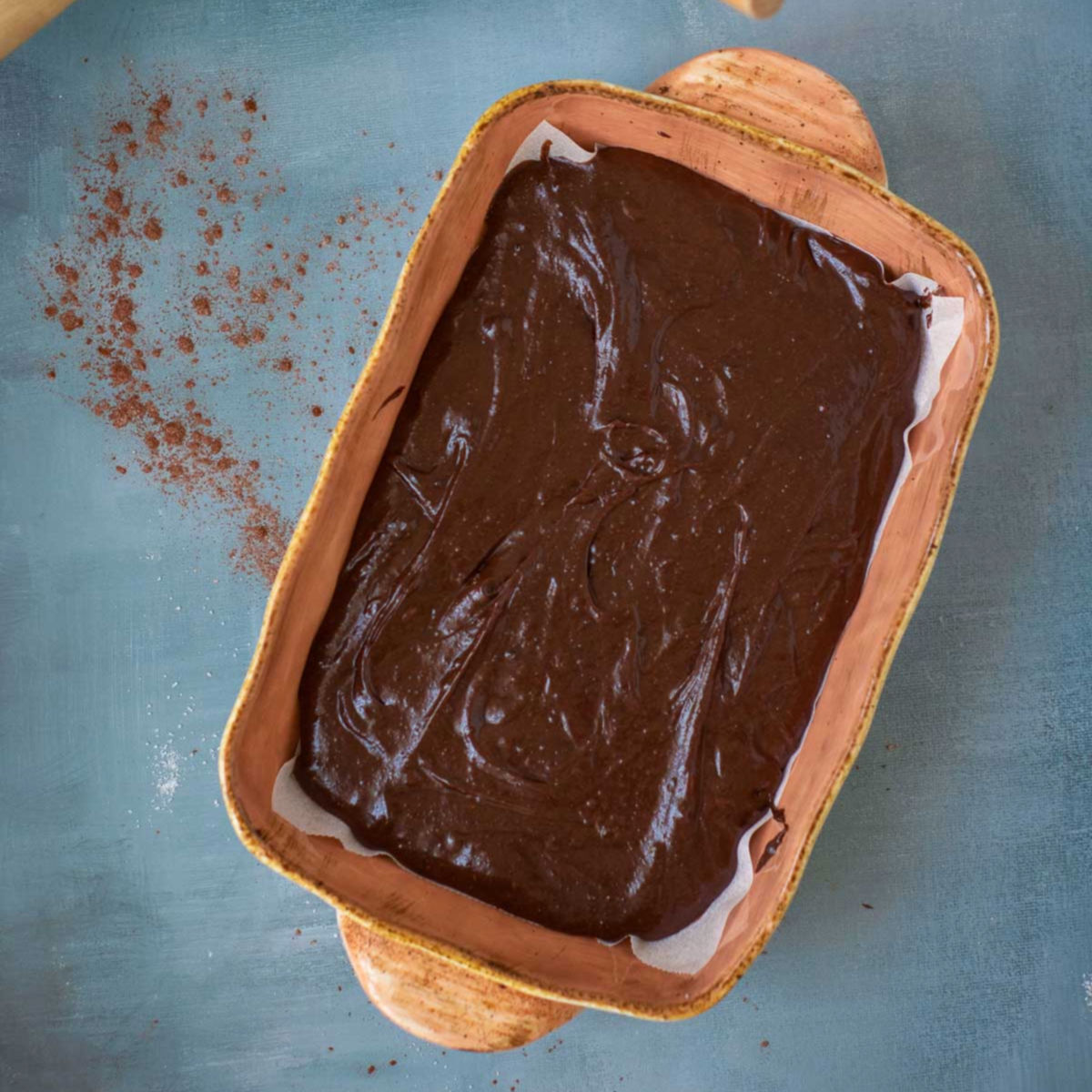 Unbaked chocolate brownie batter spread evenly in a parchment-lined rectangular baking dish.