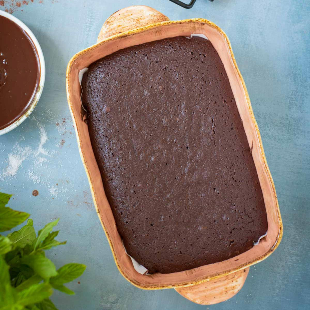 Baked brownie cake cooling in a parchment-lined baking dish on a blue surface.