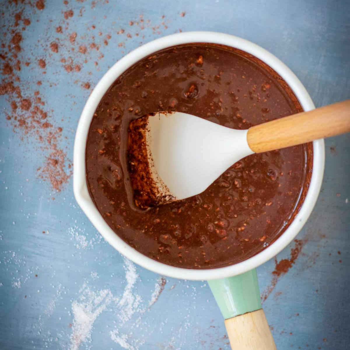 Mixing glossy chocolate ganache with a spatula in a saucepan on a blue surface.