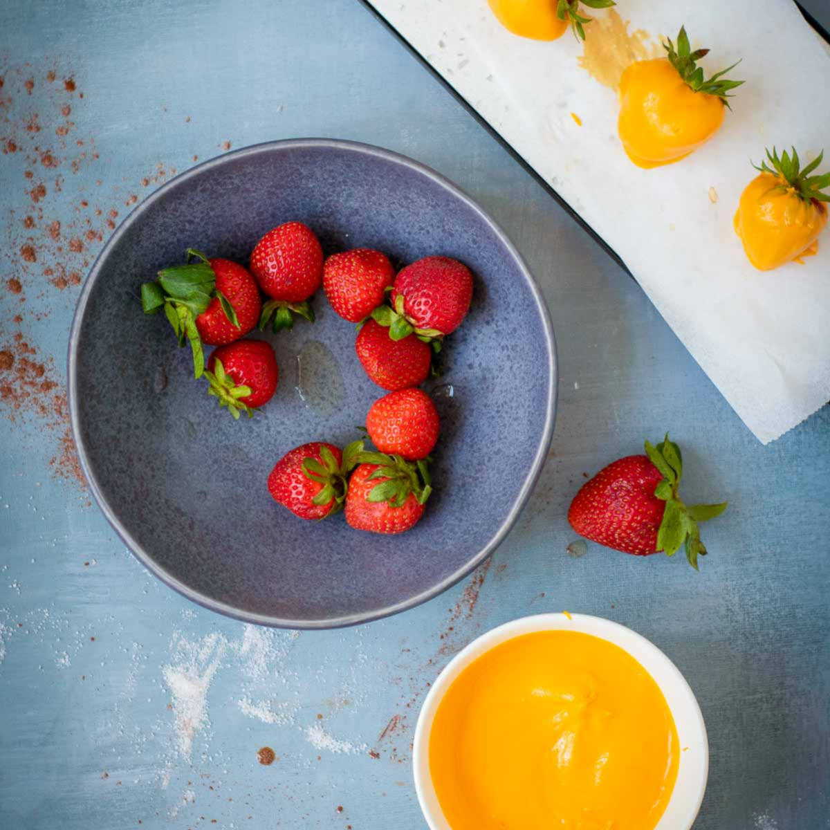 Fresh strawberries in a bowl with a dish of melted orange candy coating and dipped strawberries setting on parchment paper.