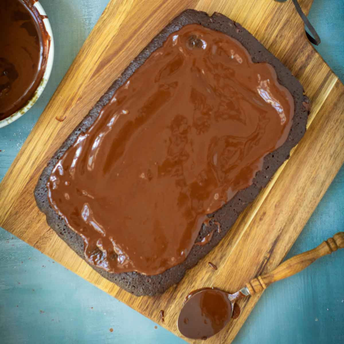 Baked brownie slab topped with smooth chocolate ganache on a wooden cutting board.