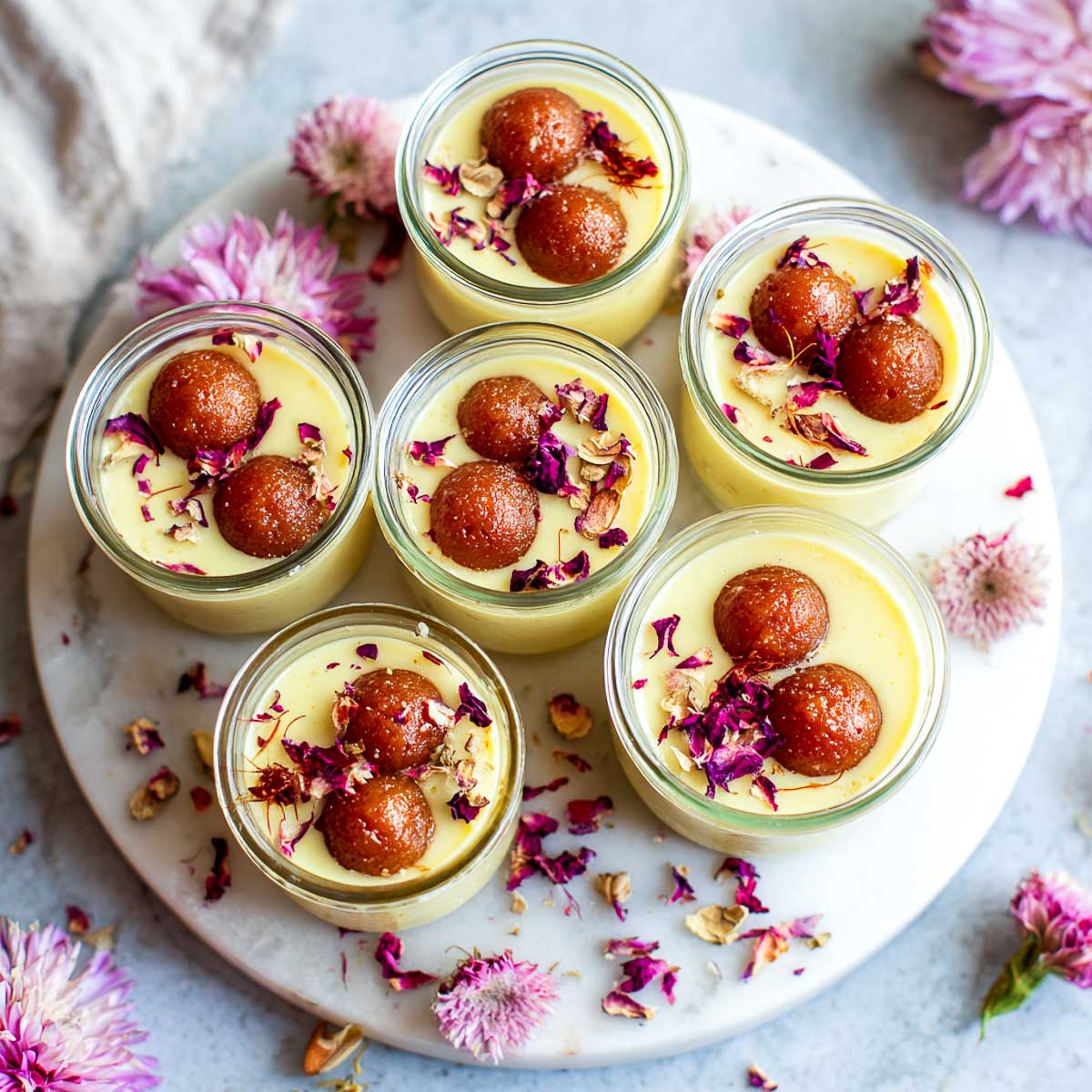 Top view of several glass jars of custard, each topped with gulab jamun and rose petals, arranged on a marble platter with flowers.
