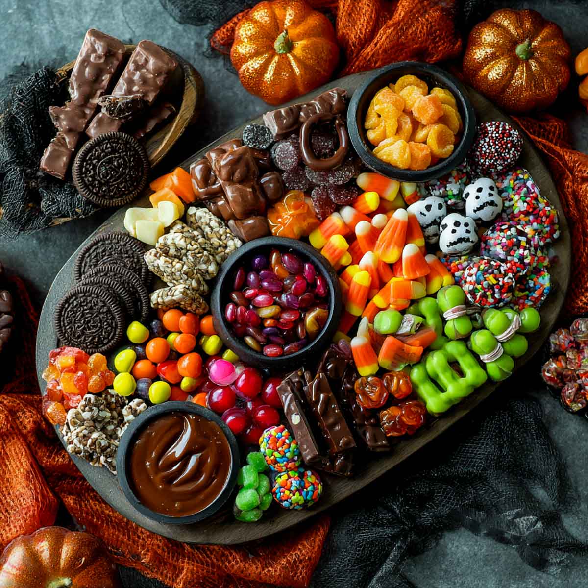 Halloween candy charcuterie board with candy corn, chocolate bars, jelly beans, ghost-shaped marshmallows, caramel dip, assorted gummies, green Halloween candies.