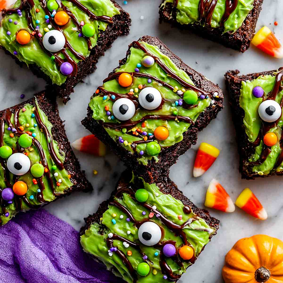 Top-down view of monster eye brownies with green frosting, candy eyeballs, colorful candies, and chocolate drizzle.