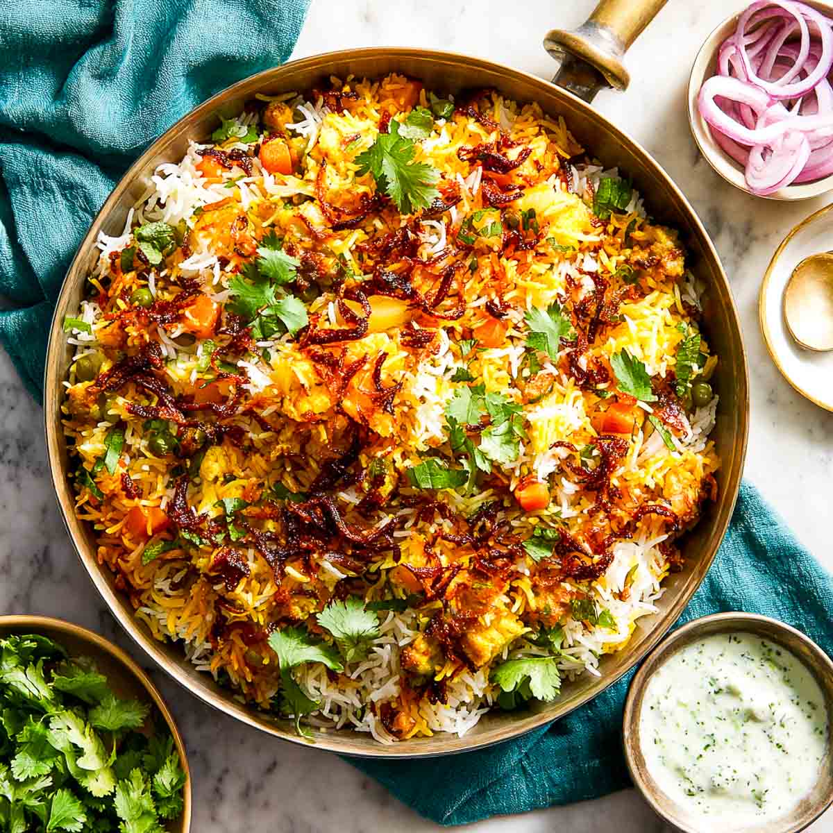 Colorful vegetable biryani in a wide pot with raita on the side.