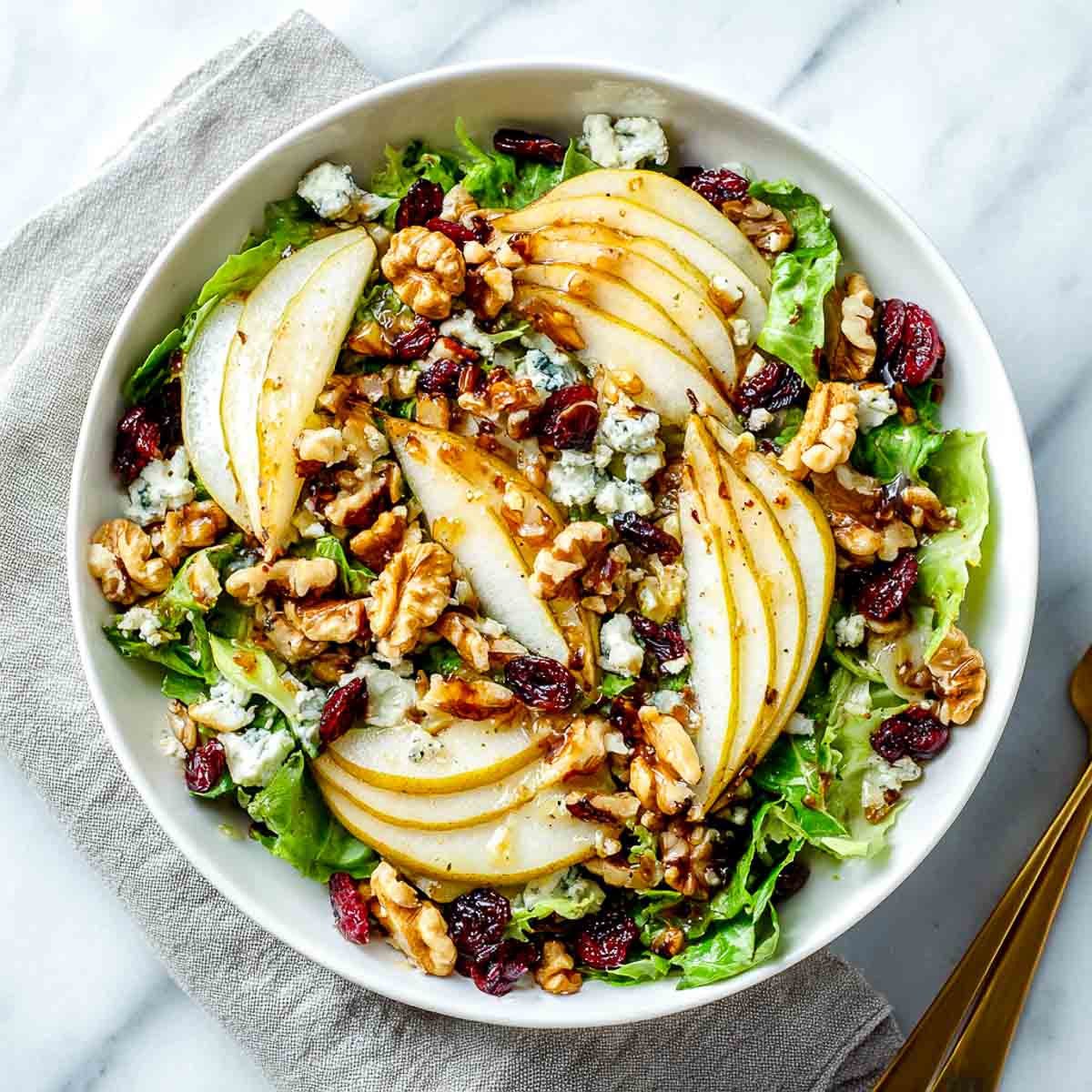 pear and walnut salad with vegan blue cheese crumbles, dried cranberries, and balsamic drizzle on fresh greens in a white bowl.