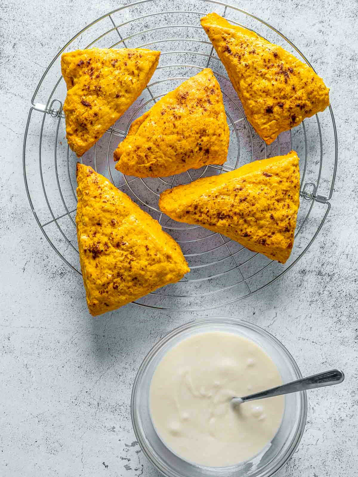 Golden pumpkin scones cooling on wire rack beside bowl of vanilla glaze.