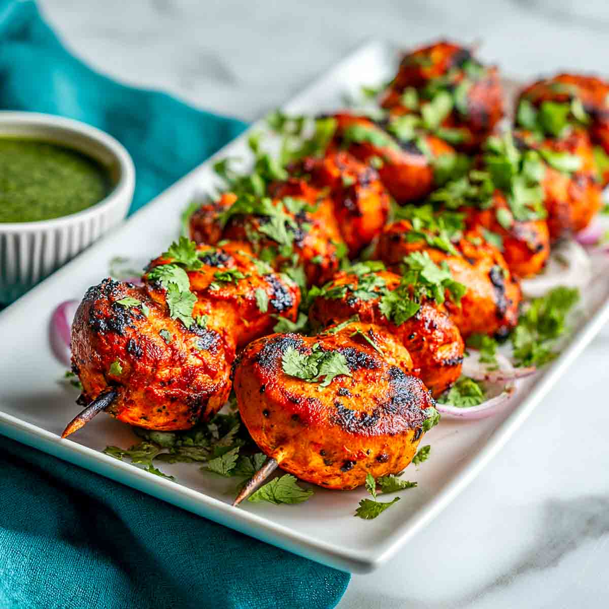 Close-up of skewered tandoori mushrooms grilled to perfection, garnished with fresh cilantro, and served on a rectangular white plate with green chutney on the side.
