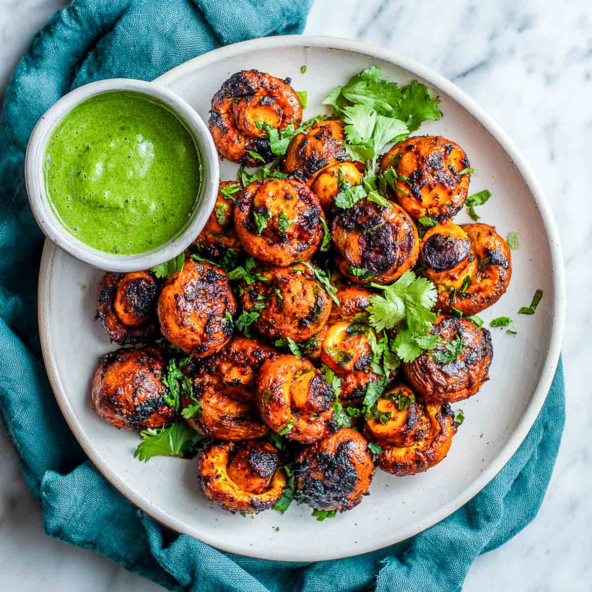 Plate of charred tandoori mushrooms garnished with cilantro, served with a bowl of green chutney, and set against a teal fabric backdrop.