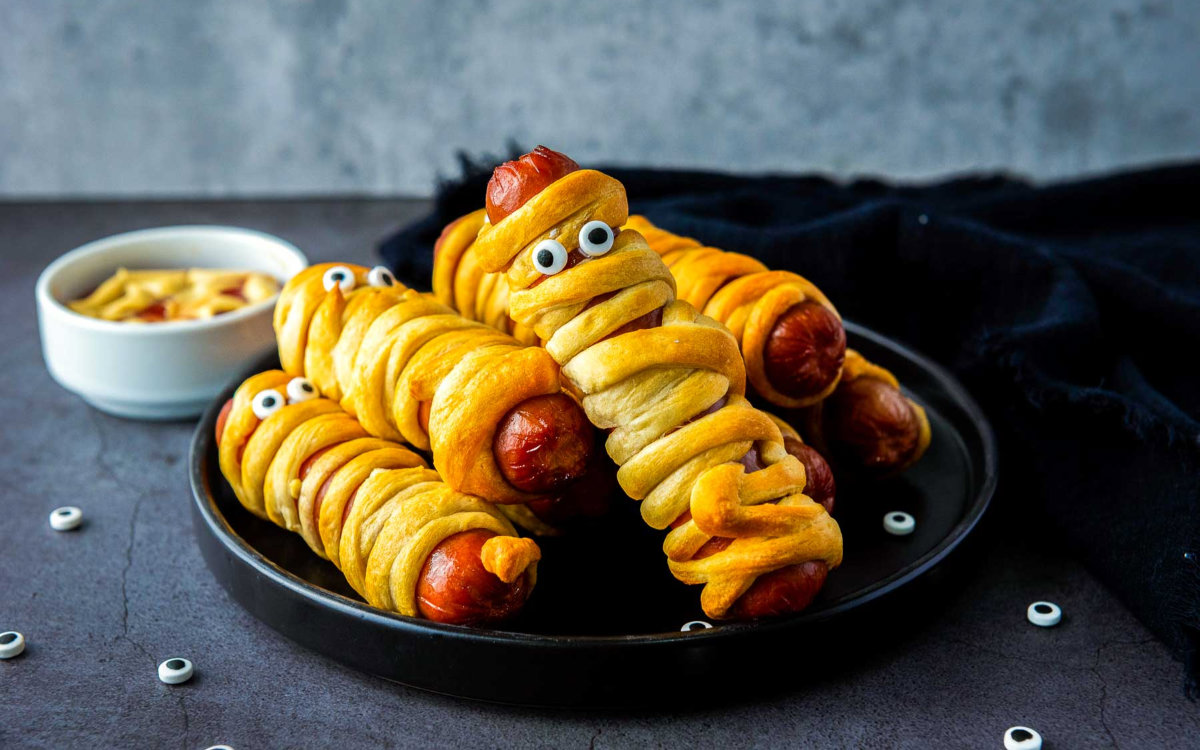 Golden brown mummy hot dogs with candy eyes served on black plate.