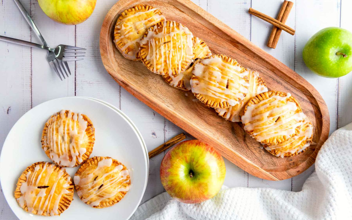 Top view of apple hand pies with icing on a wooden tray and white plate, styled with apples and cinnamon.