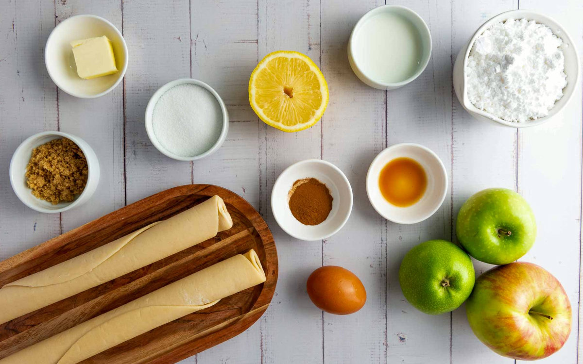 Overhead view of apple hand pie ingredients including apples, butter, sugars, cinnamon, lemon, egg, vanilla, and pie dough.