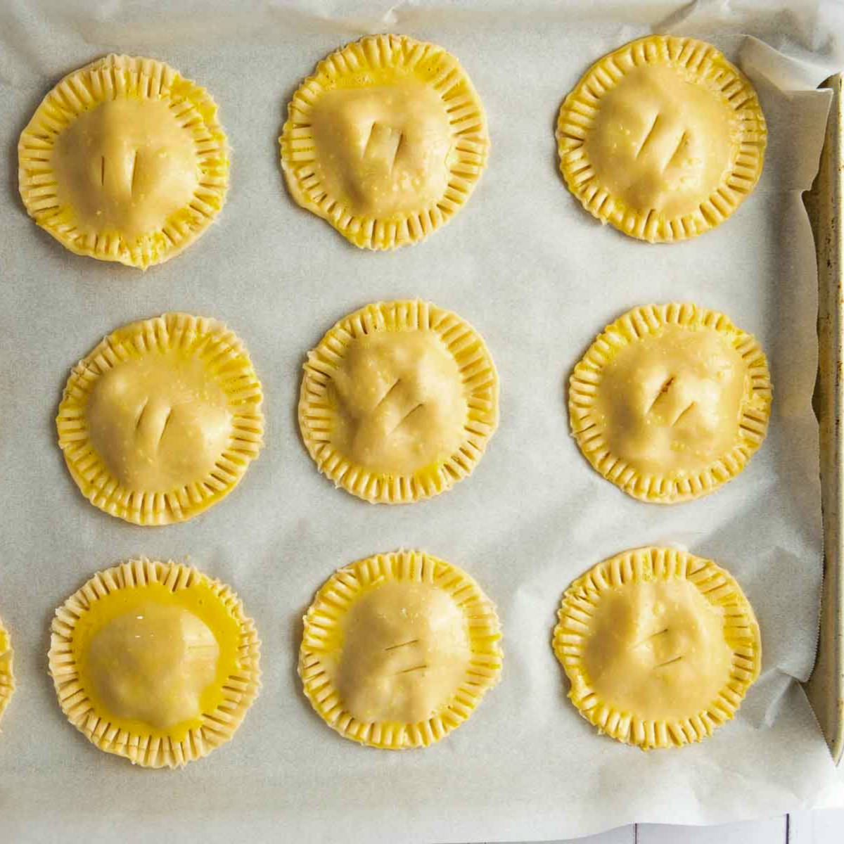 Unbaked apple hand pies sealed with fork crimping on a parchment-lined baking sheet.
