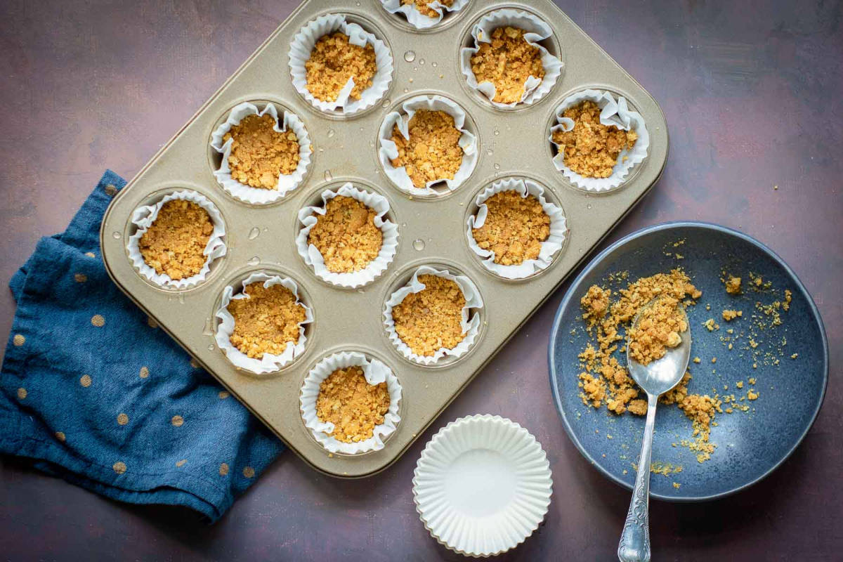 Muffin tin lined with cupcake wrappers filled with crushed graham cracker crust.