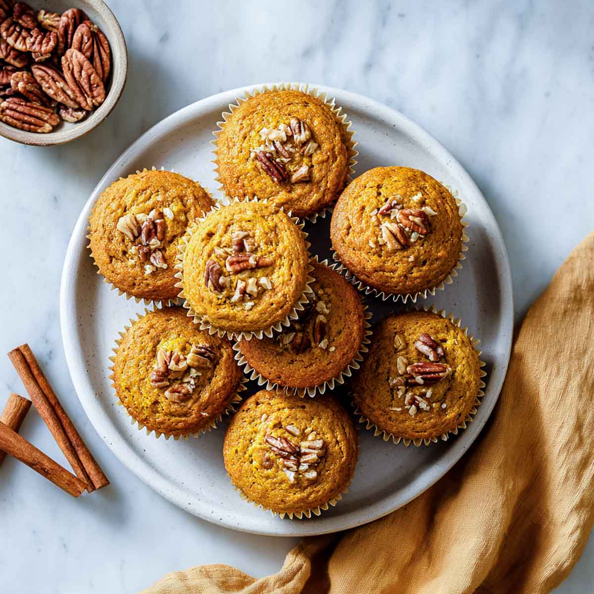 Golden keto pumpkin muffins topped with chopped pecans on white ceramic plate.